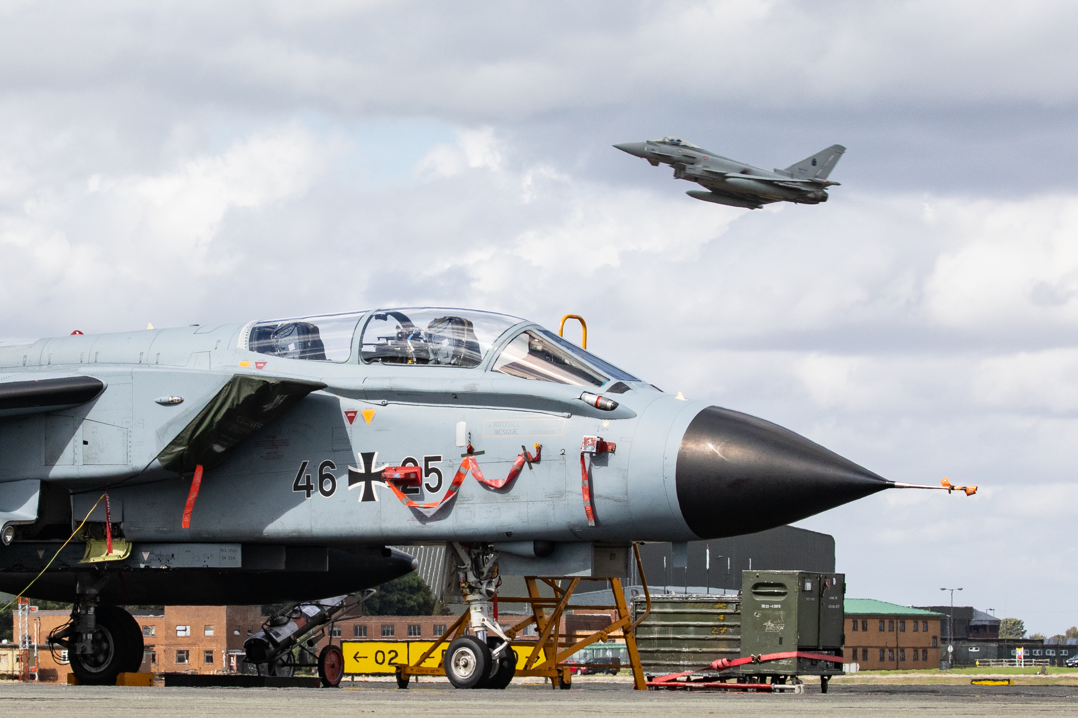 Image shows fighter jets on the runway as one flies overhead.